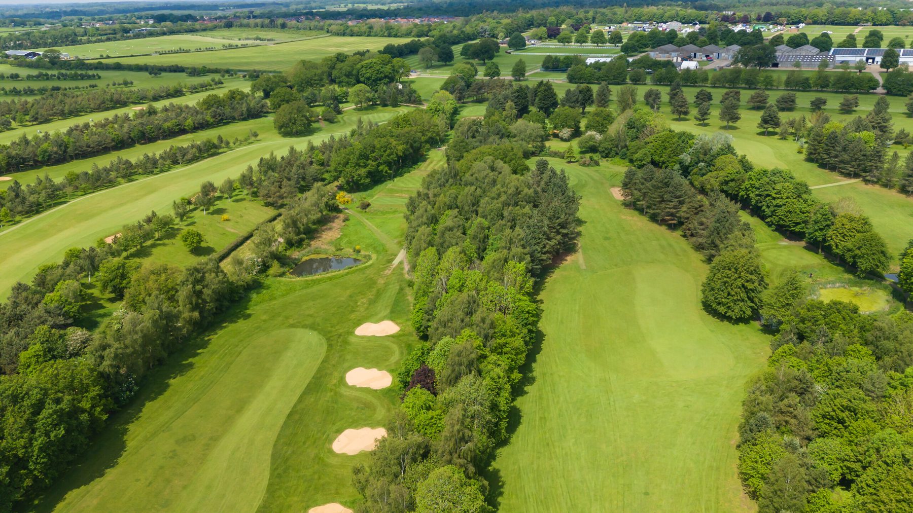 Aerial view of two parallel fairways separated by a belt of pines, with three fairway bunkers, a pond and the Norfolk Showground beyond