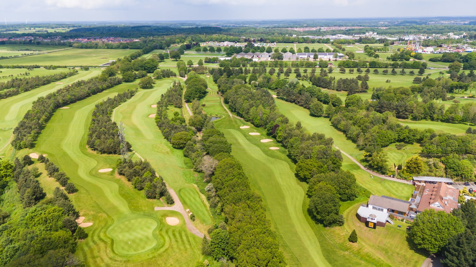 Aerial view of the course: parallel fairways divided by belts of mature trees, greens and bunkers, with the clubhouse at lower right and Norwich beyond