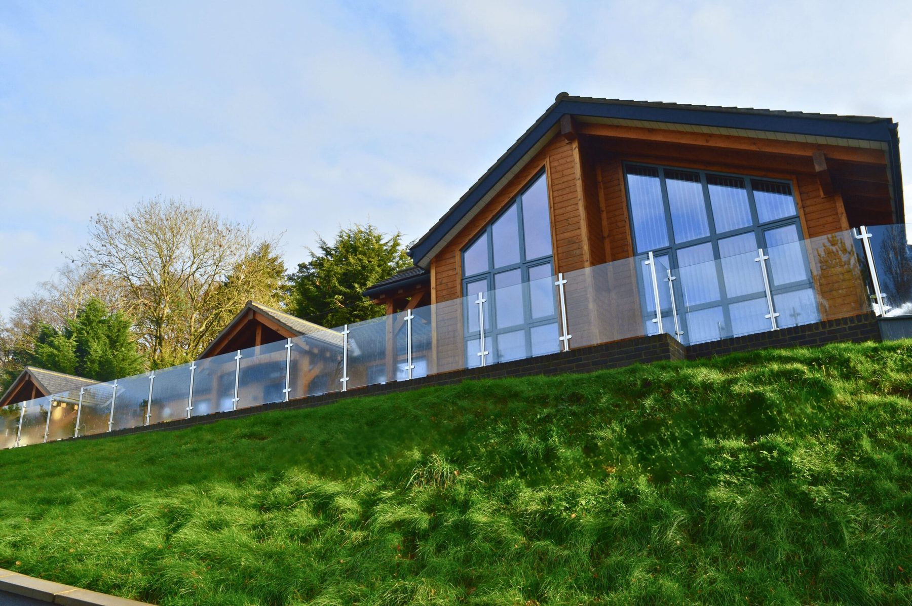 The Woodlands lodges seen from below a grass bank: timber cladding, tall glazed gables and a glass balustrade under a bright winter sky