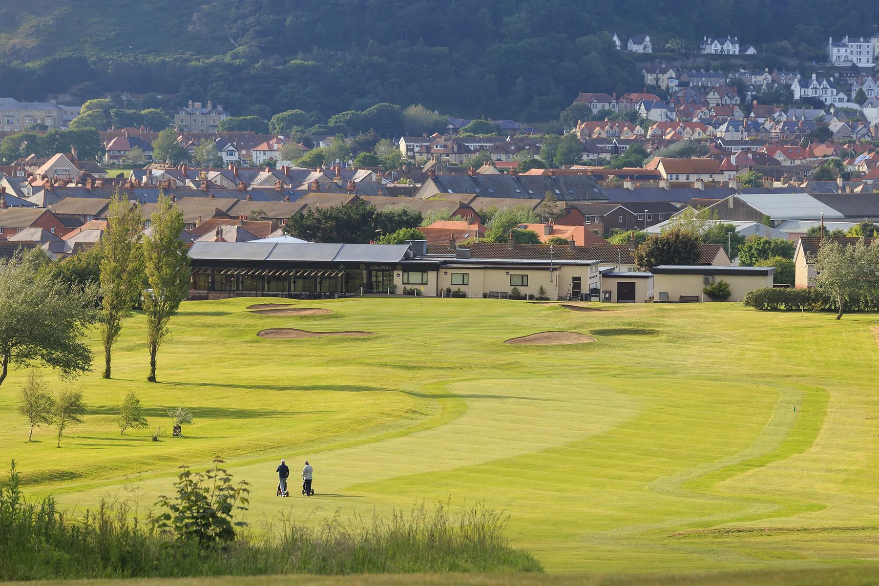 The eighteenth fairway rising towards the clubhouse, two golfers walking with trolleys, the rooftops of Llandudno and the hillside behind