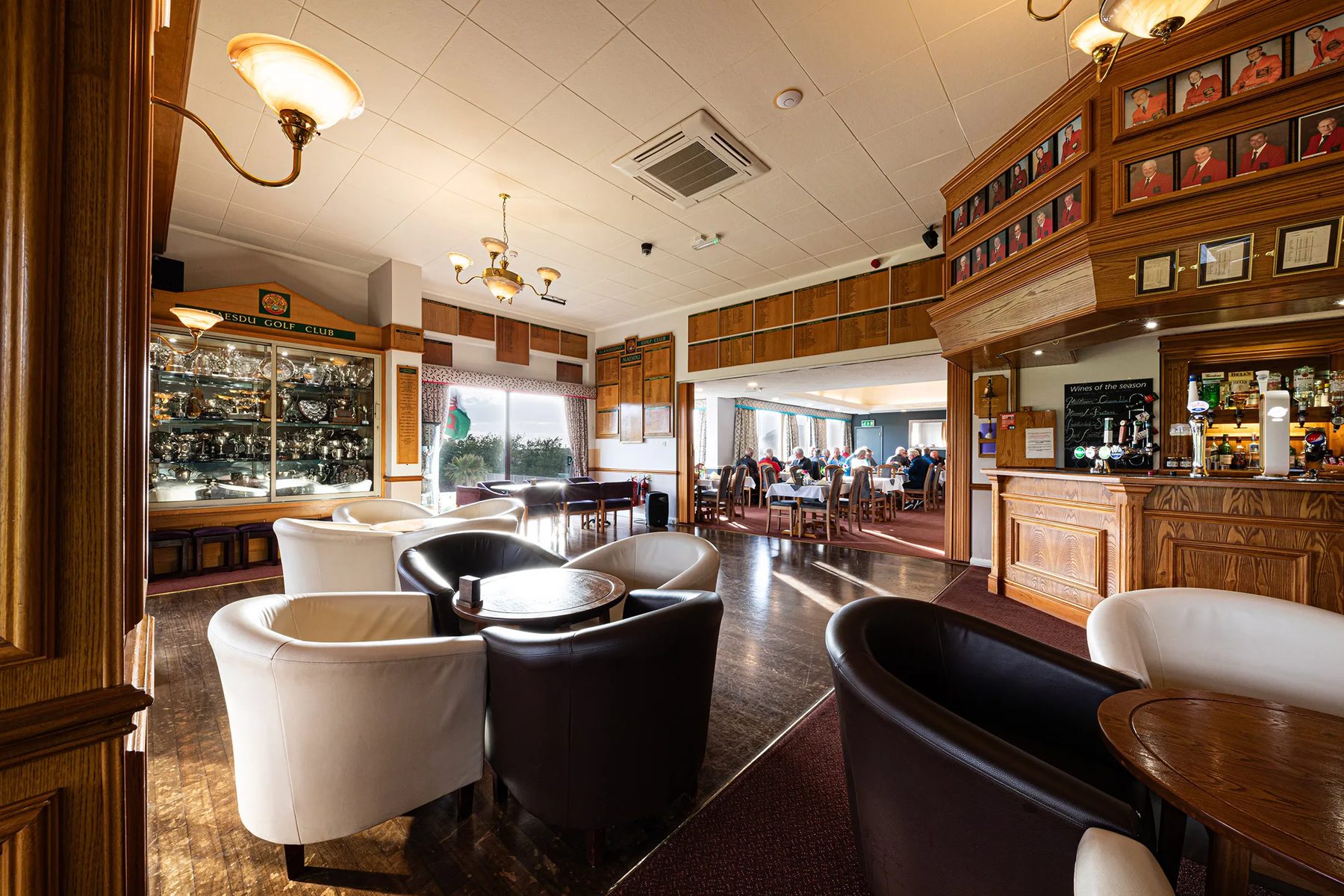 The clubhouse lounge and bar, with a trophy cabinet under a Maesdu Golf Club sign, captains' portrait boards on the right and diners in the room beyond