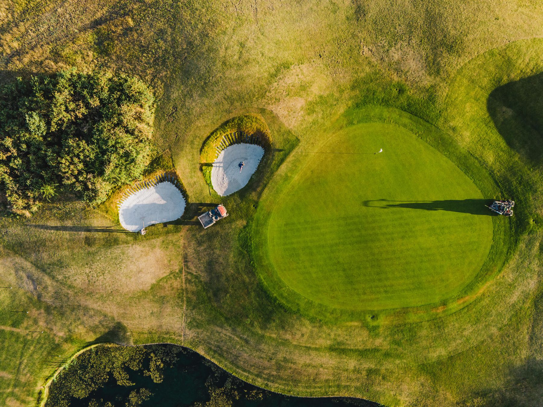 Straight-down aerial of a green and two revetted bunkers, a greenkeeper raking sand beside a utility vehicle, long evening shadows across the turf