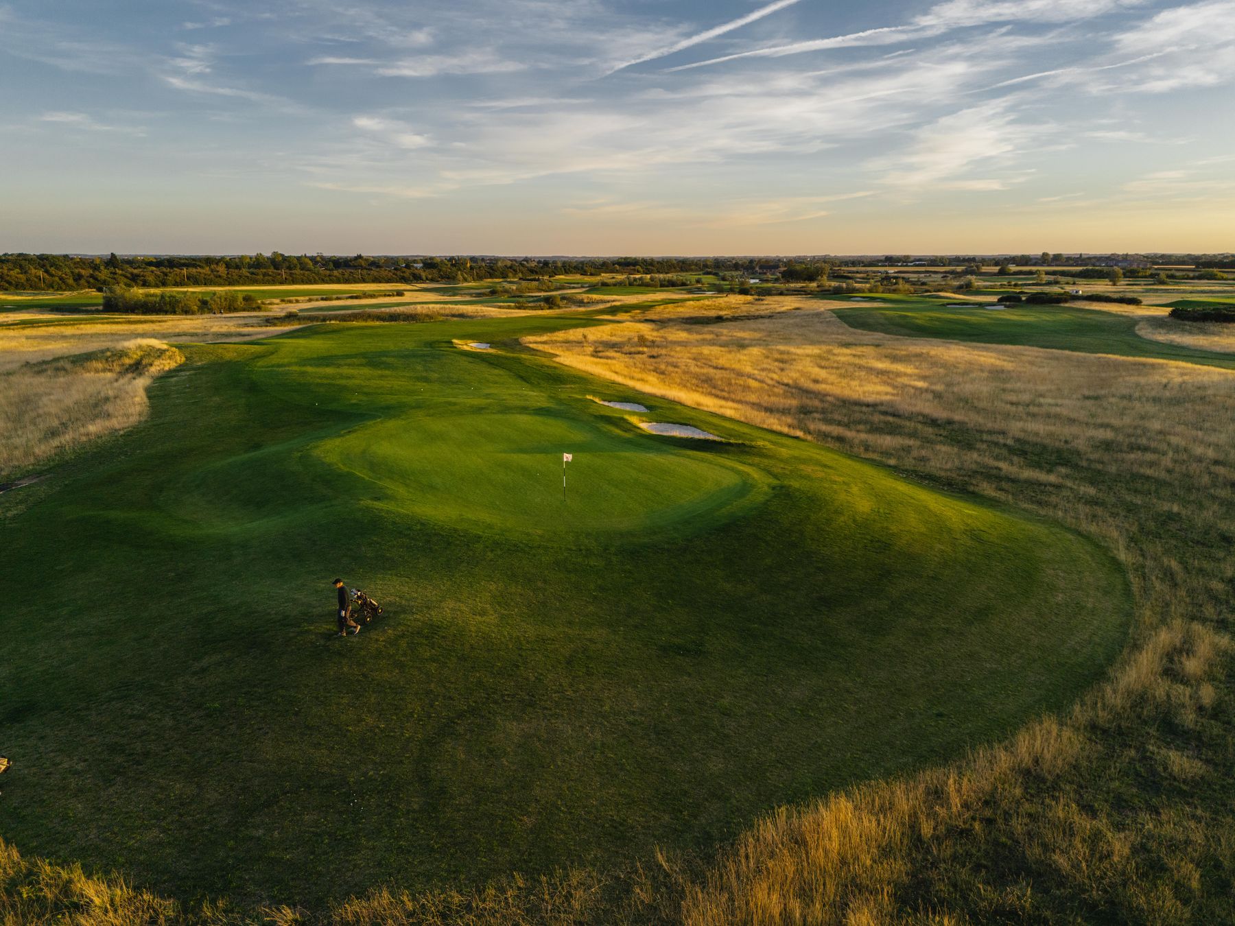 Golden-hour aerial of a green guarded by three revetted bunkers, a lone golfer pulling a trolley across the fairway, the rough turning gold