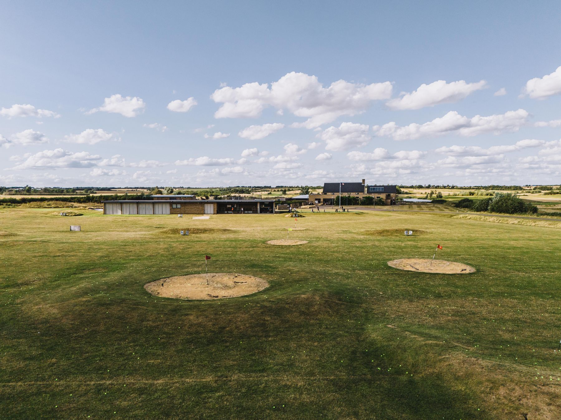 Aerial view over the practice ground to the range building and clubhouse, target greens cut into the grass, flat Fenland to the horizon under a cloud-scattered sky