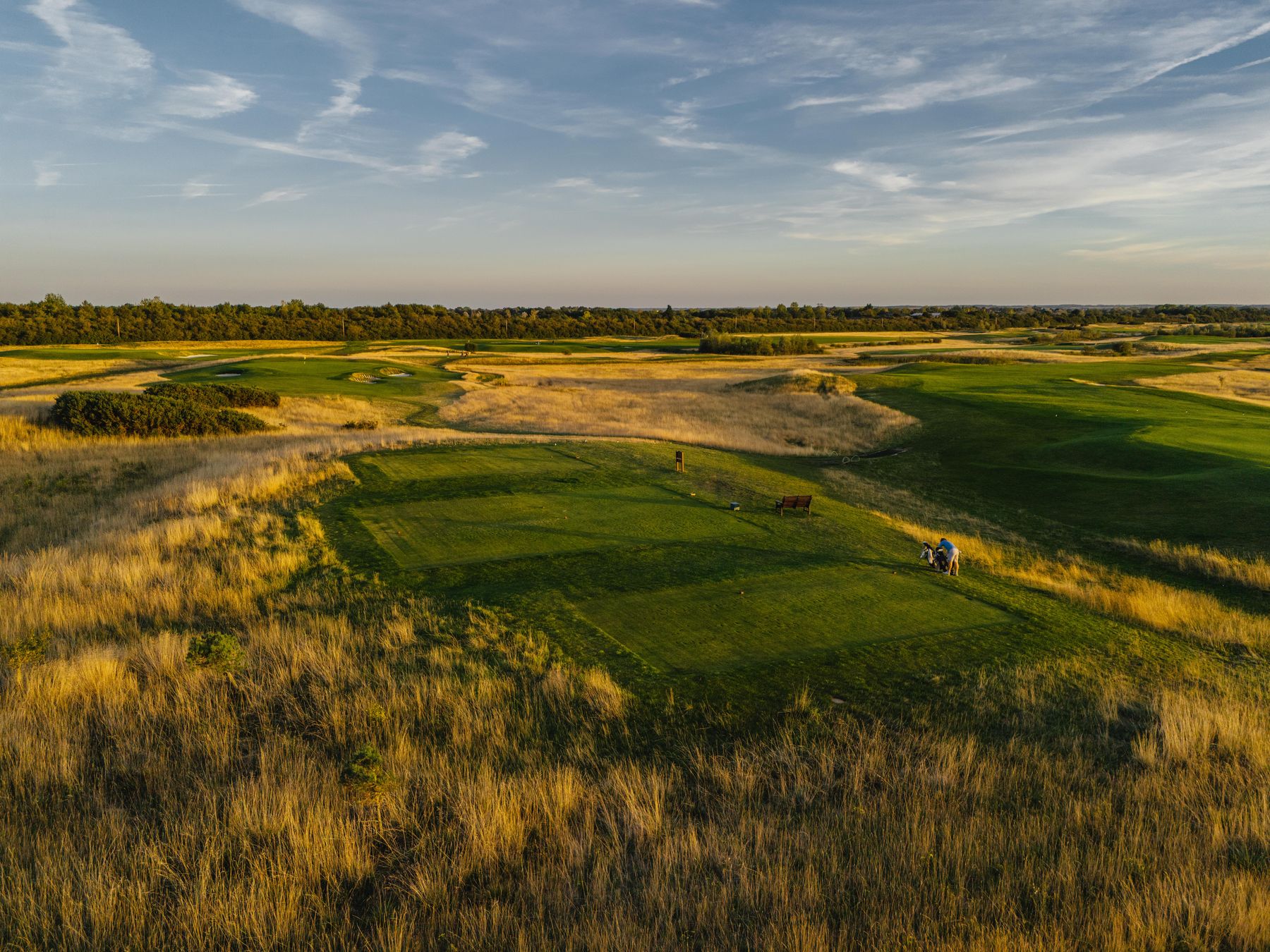 Golden-hour aerial of a tee cut into fescue, two golfers with a trolley waiting to play, the course running to a low tree line under a wide sky