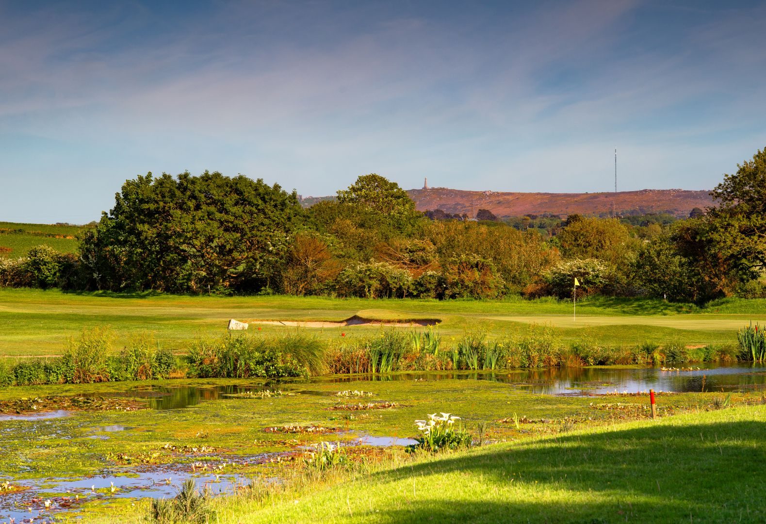 A lake thick with water lilies in the foreground, a bunkered green with a yellow flag beyond it, and the Carn Brea monument and a mast on the ridge behind